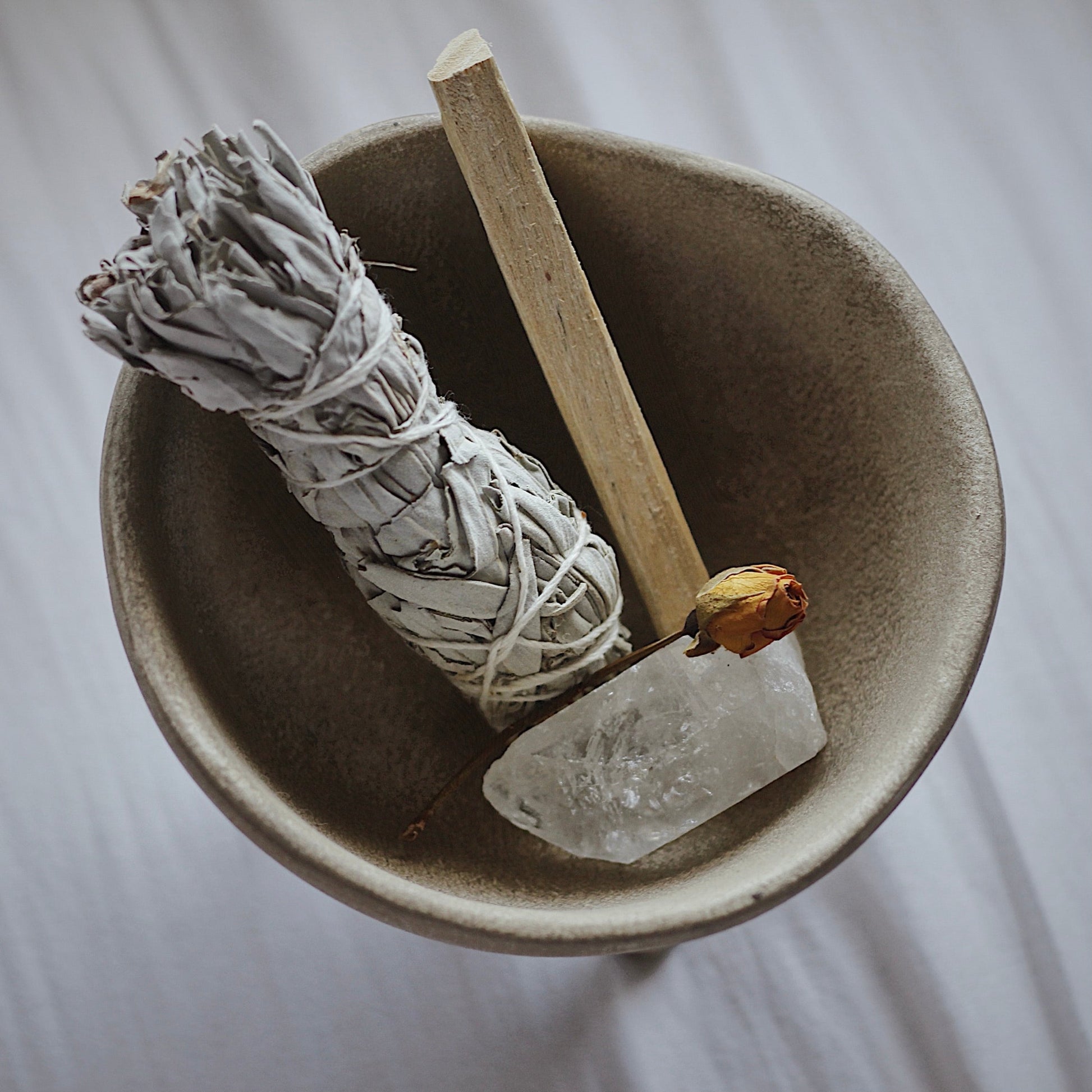 A photo showing a light brown ceramic bowl, contains a white sage stick, a crystal, a dried flower bud, to use in the ritual of smudging.